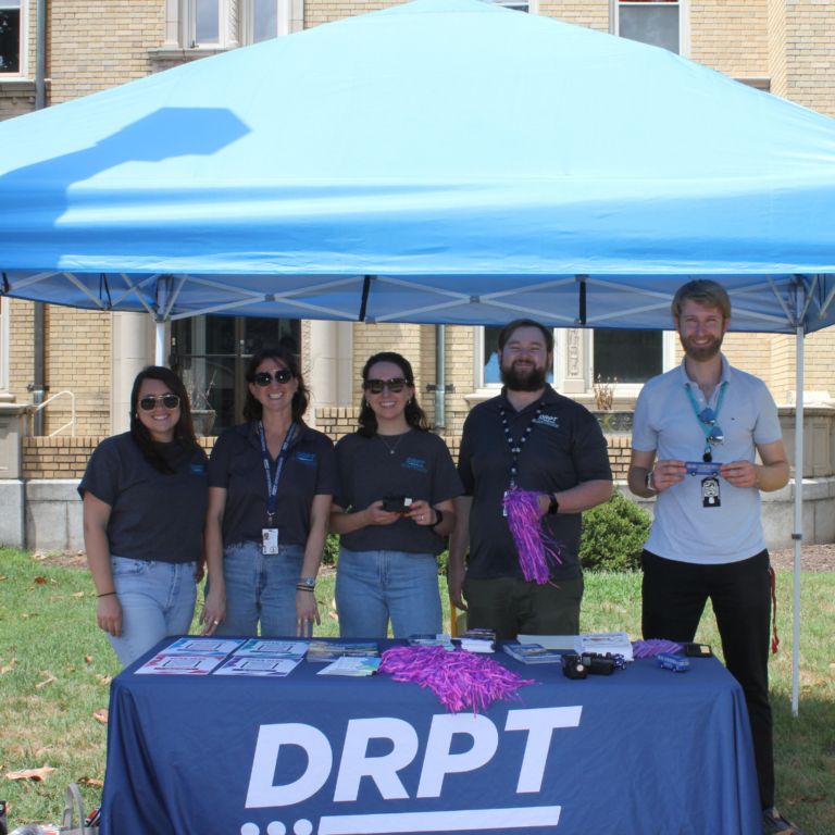El personal de DRPT sostiene artículos promocionales en una mesa de la marca DRPT bajo una carpa azul