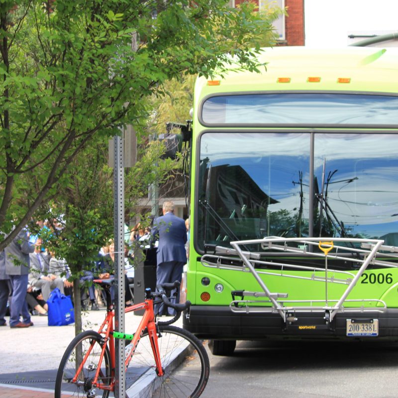 Una imagen fotográfica de un autobús aparcado junto a una acera abarrotada con peatones al fondo y una bicicleta en primer plano.